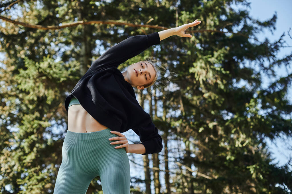 подходит молодая женщина с светлыми волосами и спортивной одеждой растягивая свое тело во время тренировки в парке - Фото, изображение