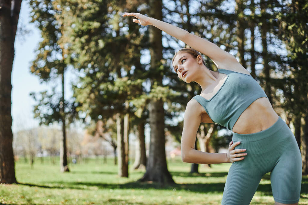 young woman with blonde hair and sportswear stretching her body while putting hand on waist in park - Photo, Image