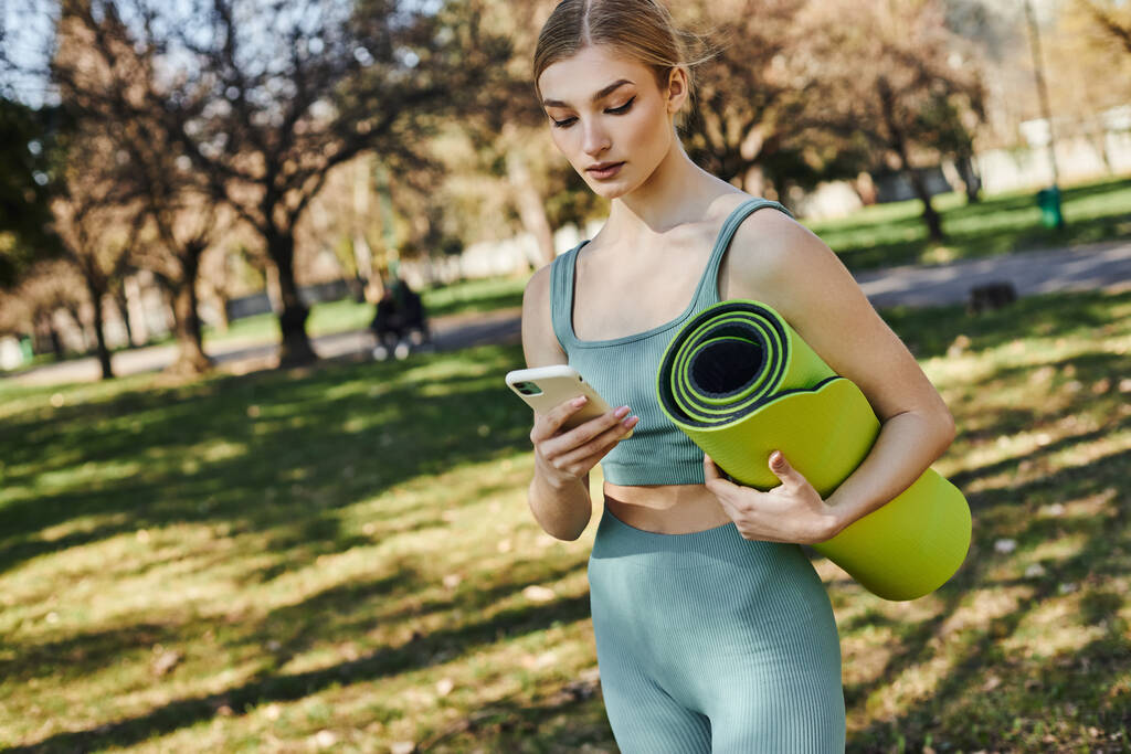Aktif giyinen güzel sporcu kadın akıllı telefon kullanıyor ve parkta dururken spor minderi tutuyor. - Fotoğraf, Görsel