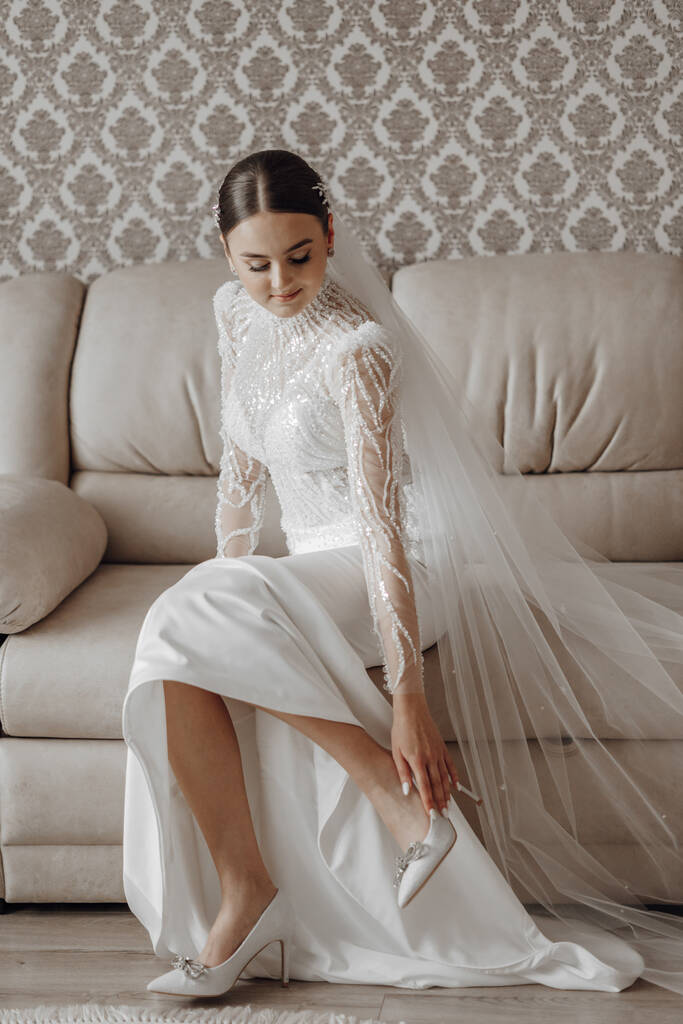 Невеста в свадебном платье надевает обувь на ноги, сидя на диване в своей комнате - Фото, изображение