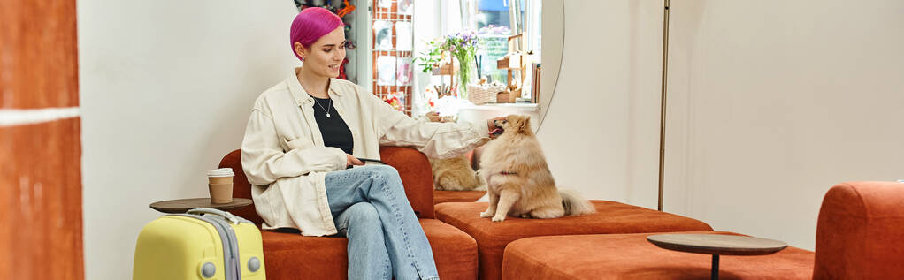 Schicke Frau mit Smartphone kuschelt Pommernspitz neben Reisetasche im Aufenthaltsraum des Haustierhotels - Foto, Bild