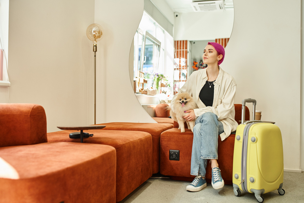 lila hajú nő, pomerániai köpettel és bőrönddel a kedvtelésből tartott szálloda halljában - Fotó, kép