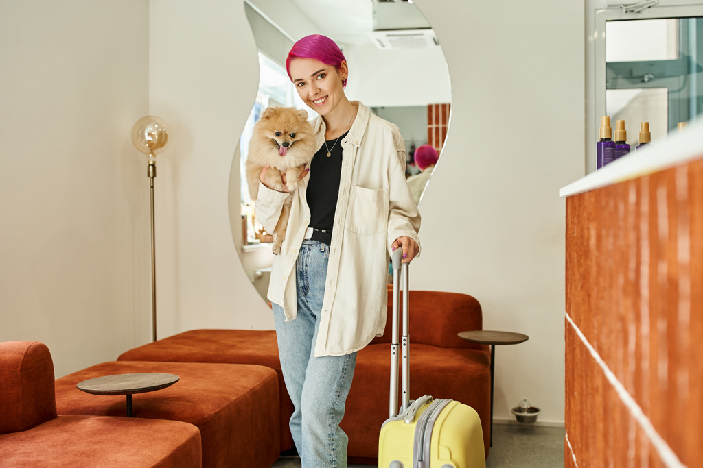 alegre mujer de pelo morado mirando a la cámara mientras está de pie con perro peludo y maleta en el hotel de mascotas - Foto, imagen