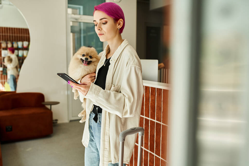 woman with pomeranian spitz messaging on smartphone near reception desk and suitcase in dog hotel - Photo, Image