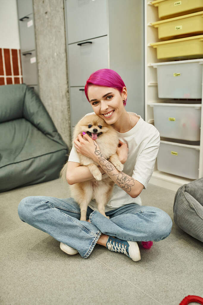 татуйована няня з фіолетовим волоссям, що обіймає пухнастого собаку і посміхається на камеру на підлозі в готелі - Фото, зображення