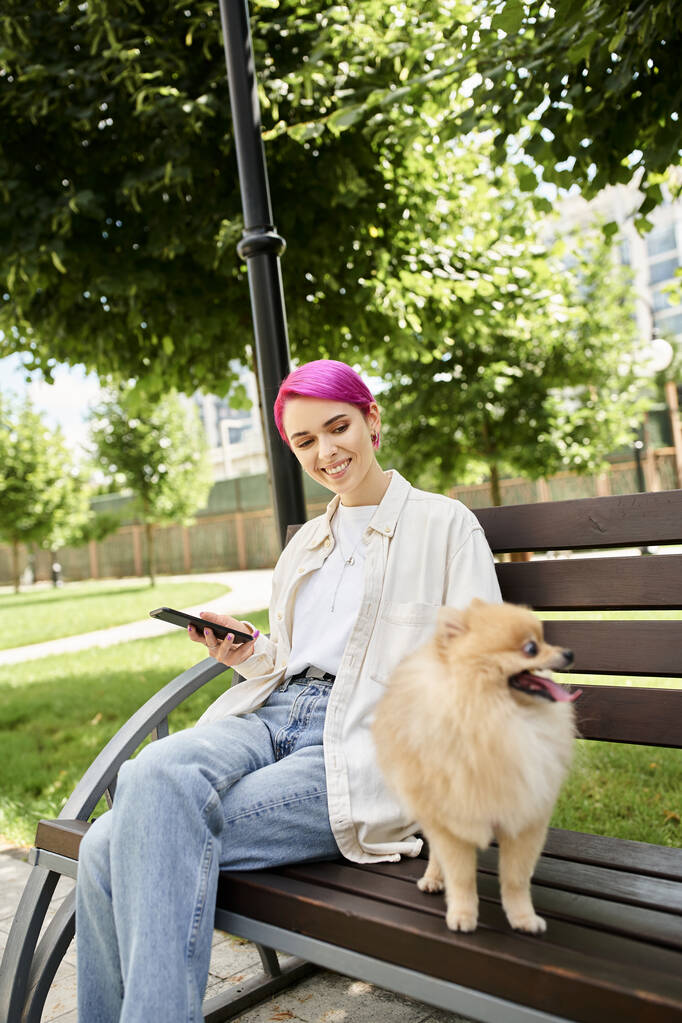 cheerful purple-haired woman with smartphone looking at loveable pomeranian spitz on bench in park - Photo, Image