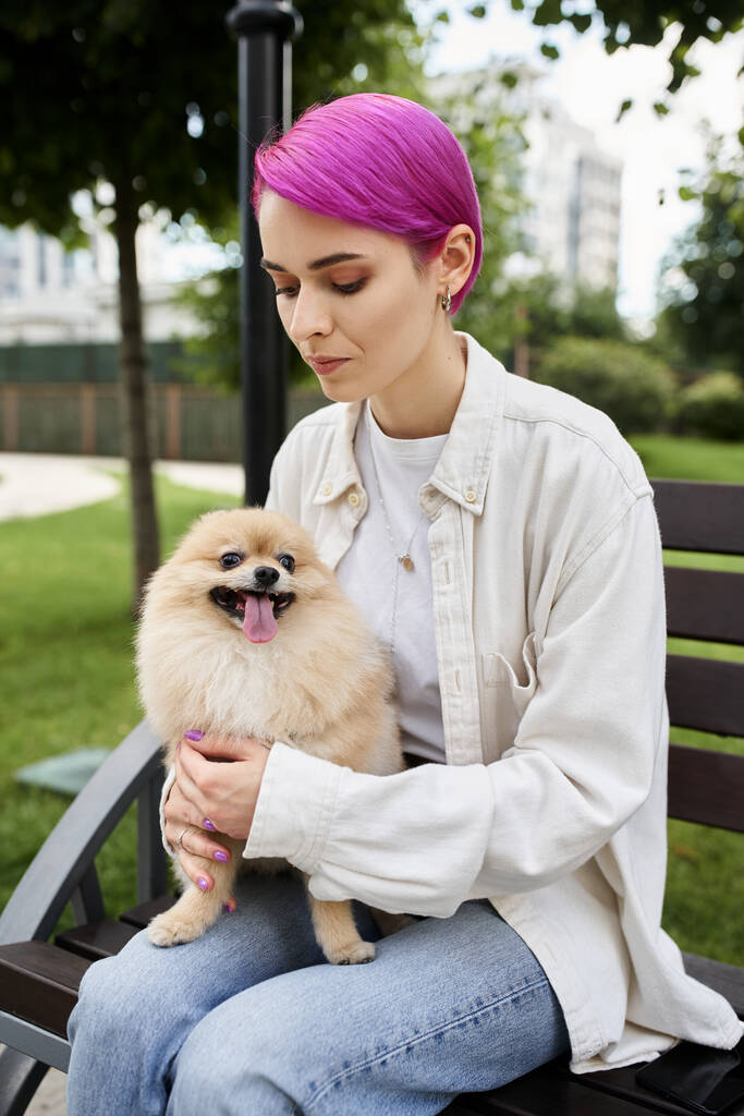 стильна фіолетово-волохата жінка, що обіймає португальський шпіц, сидячи на лавці в парку - Фото, зображення