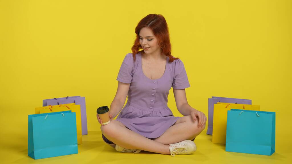 Een roodharige vrouw zit gekruist op de vloer, met een papieren kopje koffie of thee. Jonge vrouw in zomerkleren omgeven door kleurrijke boodschappentassen geïsoleerd op gele achtergrond. Concept van - Foto, afbeelding