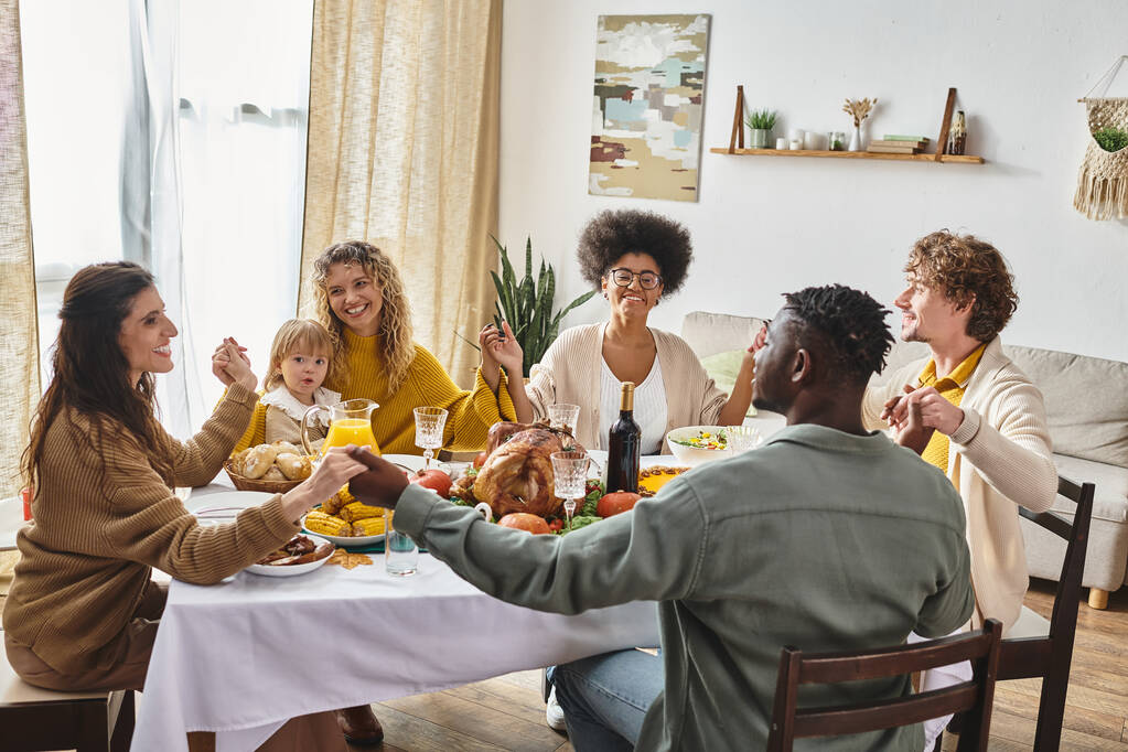 szczęśliwy wielorasowych przyjaciół i rodziny trzymając się za ręce i modląc się przy stole Dziękczynienia, wdzięczny - Zdjęcie, obraz