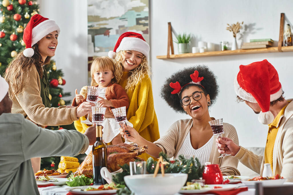 大きい多民族の喜びの家族はサンタの帽子を身に着けているお祝いのテーブルでメガネをかぶっています,クリスマス - 写真・画像