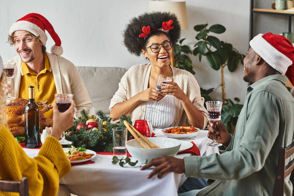 gran familia multicultural hablando y sonriendo alegremente en la mesa de Navidad con sombreros de Santa - Foto, imagen