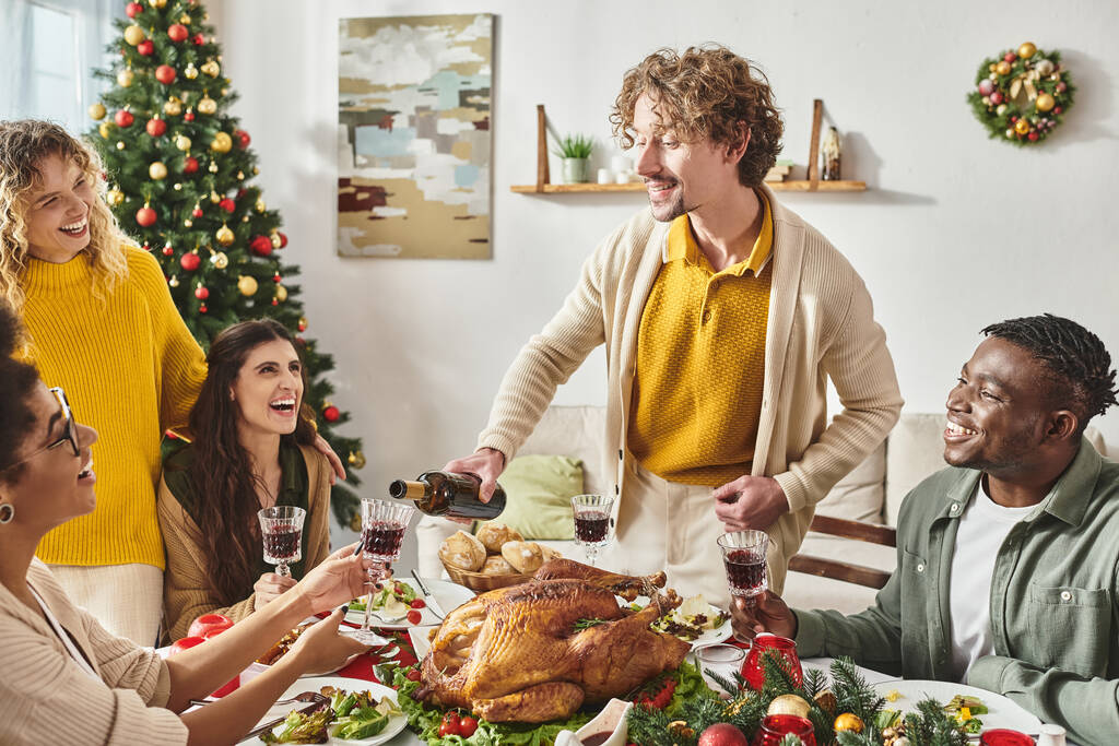 giovane uomo sorridente circondato dai suoi parenti versando del vino in un bicchiere vicino all'albero di Natale - Foto, immagini