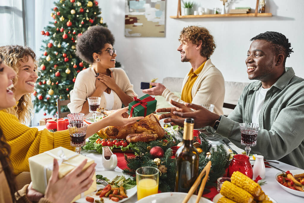 Büyük, çok kültürlü bir aile şenlik masasında oturmuş Noel ağacıyla hediye alışverişinde bulunuyorlar. - Fotoğraf, Görsel
