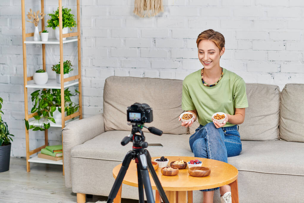 femme végétarienne heureuse avec des noix présentant ensemble d'ingrédients à base de plantes pendant blog vidéo à la maison - Photo, image