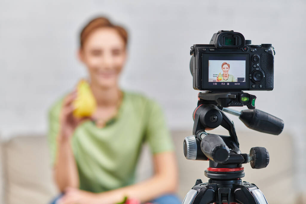 foco seletivo na câmera digital perto de mulher desfocada com cais maduro, vídeo blog on plant-based diet - Foto, Imagem