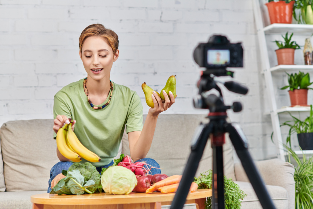 nuori nainen, jolla on kypsiä banaaneja lähellä erilaisia hedelmiä ja vihanneksia hämärtyneen digitaalikameran edessä - Valokuva, kuva