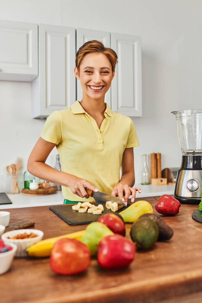 счастливая вегетарианка режет спелый банан рядом со свежими фруктами и электрическим блендером на современной кухне - Фото, изображение