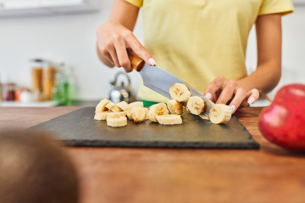vista parcial de la joven mujer cortando plátano maduro mientras cocina comida vegetariana en la cocina en casa - Foto, Imagen