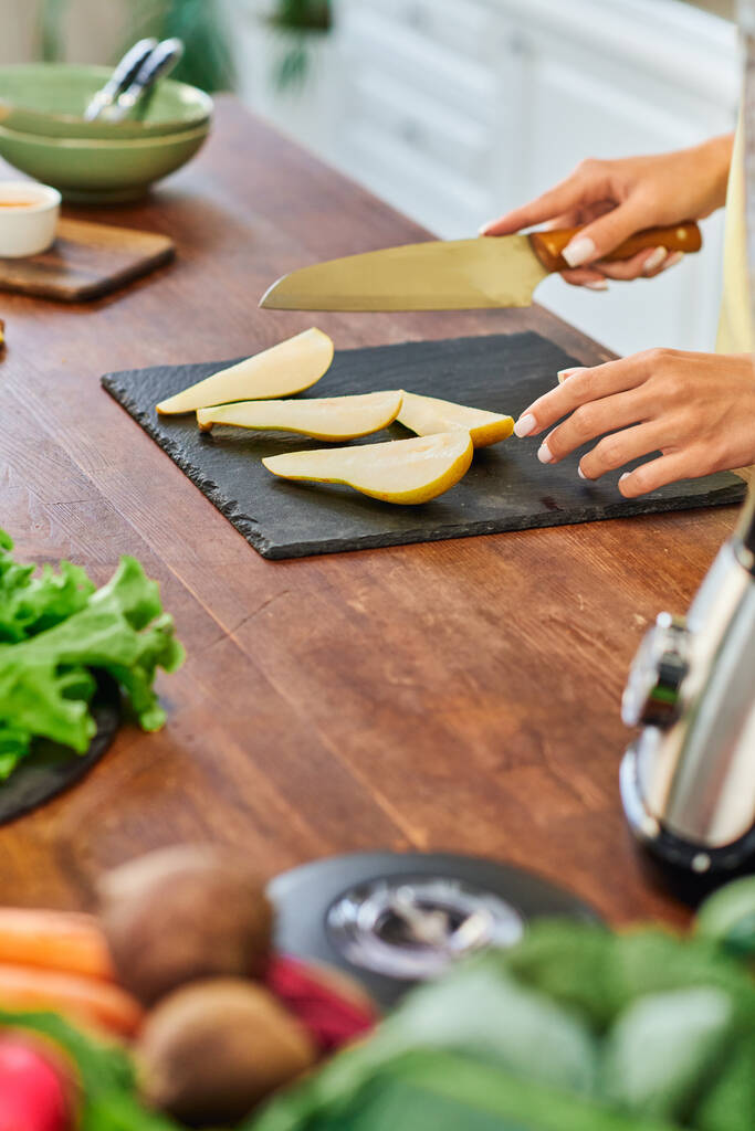 キッチンでチョッピングボード上の熟したスライスされた梨の近くのナイフを持つベジタリアン女性の部分的な眺め - 写真・画像