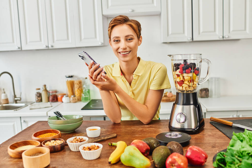 mulher vegetariana com smartphone sorrindo para a câmera perto de alimentos de origem vegetal e liquidificador com frutas - Foto, Imagem