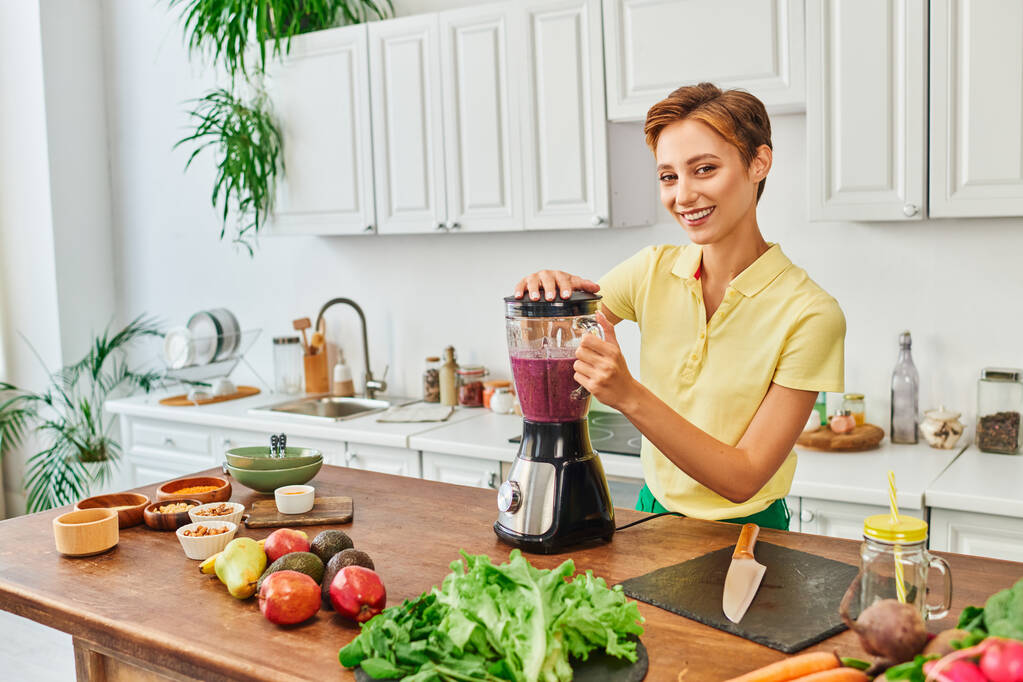 vegetarische vrouw het bereiden van smoothie in elektrische blender in de buurt van vers fruit en groenten in de keuken - Foto, afbeelding