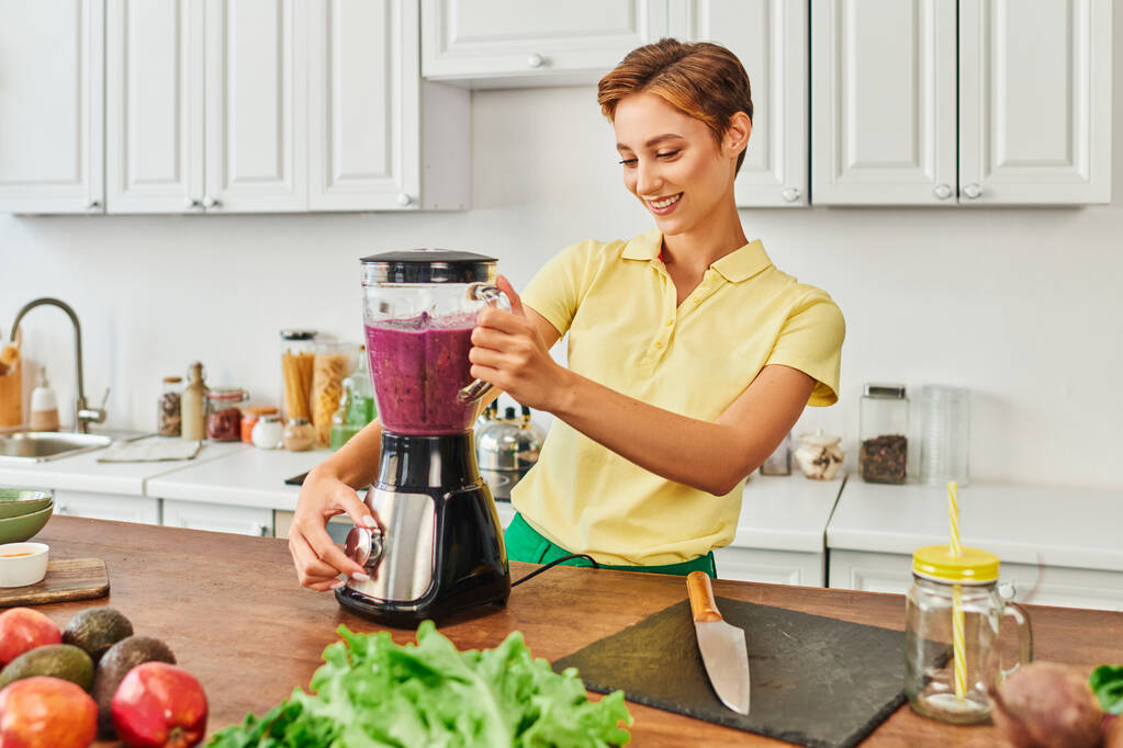 Smiley-Frau bereitet vegetarischen Smoothie im elektrischen Mixer in der Nähe von frischem Obst und Gemüse zu - Foto, Bild
