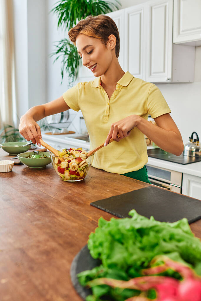 молодая и радостная женщина смешивает свежий фруктовый салат во время приготовления вегетарианской еды на современной кухне - Фото, изображение