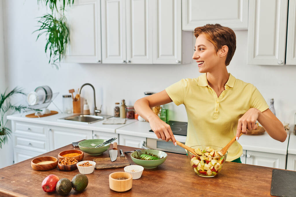 donna allegra mescolando insalata vegetariana portare frutta fresca e guardando altrove in cucina moderna - Foto, immagini
