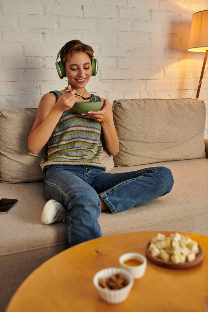 femme joyeuse dans les écouteurs ayant un dîner végétarien près sur le canapé près de la table avec un repas à base de plantes - Photo, image