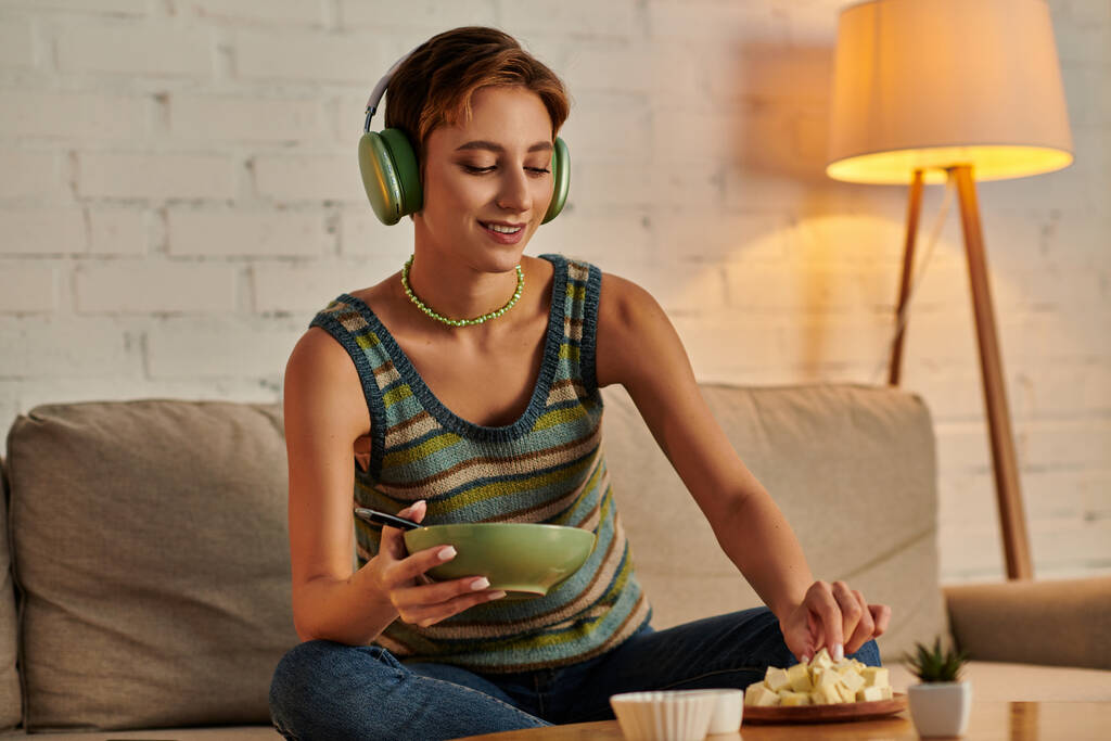 Smiley-Frau mit Kopfhörer hält Salatschüssel und nimmt Tofu-Käse, vegetarisches Abendessen auf der Couch - Foto, Bild