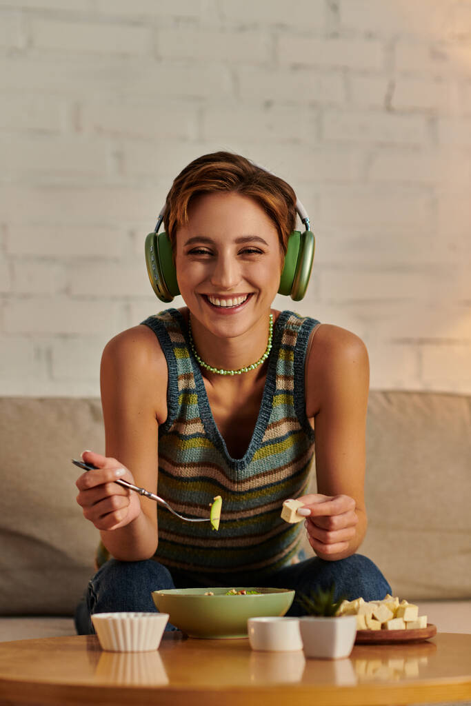 весела весела жінка в навушниках дивиться на камеру біля салата на рослинній основі і сиру тофу - Фото, зображення