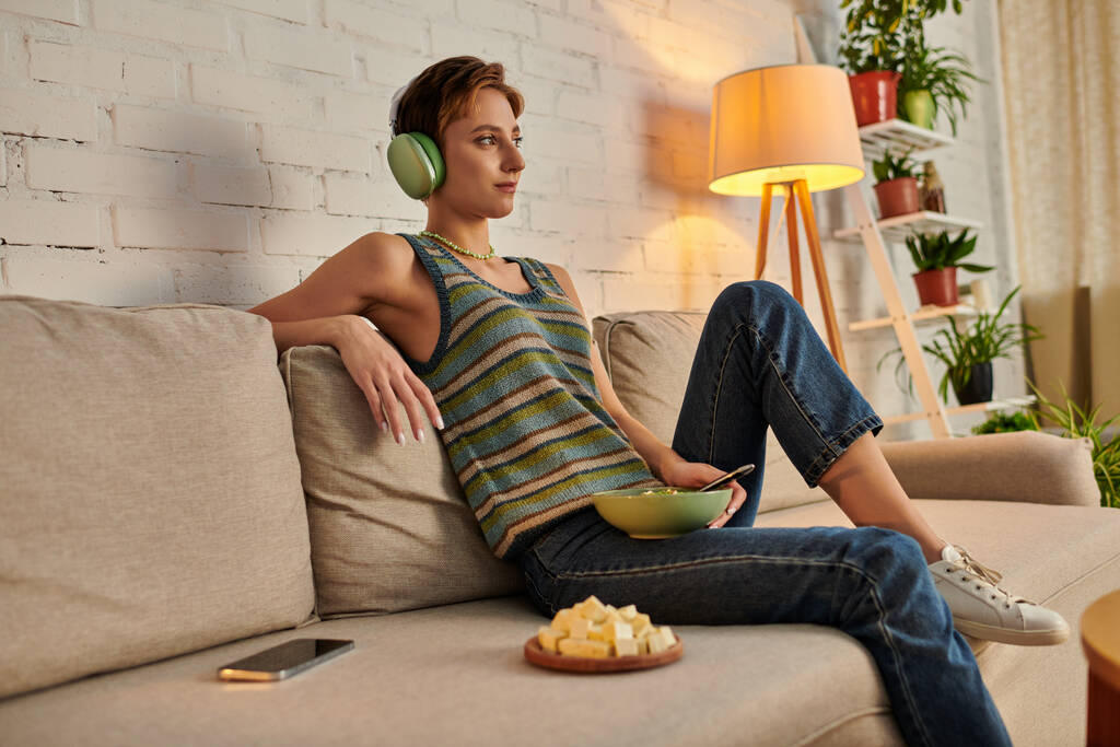 mujer vegetariana de ensueño en auriculares sentados con ensaladera cerca de queso tofu y teléfono inteligente en el sofá - Foto, imagen