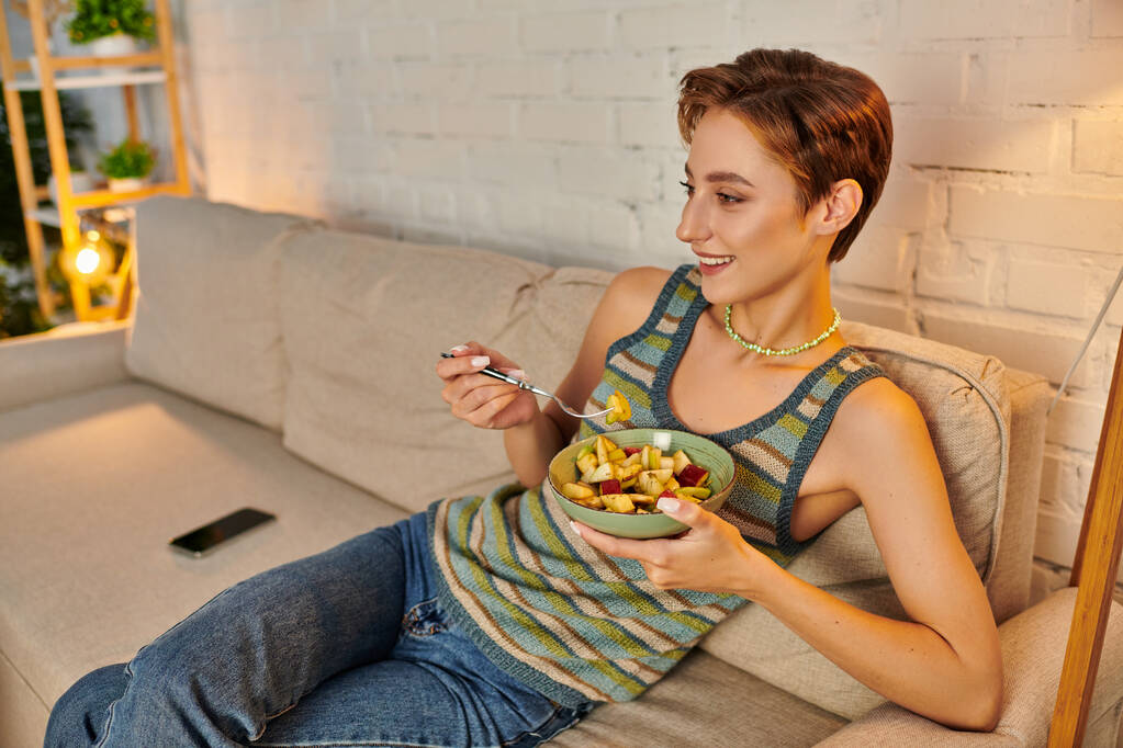 χαμογελαστή γυναίκα κάθεται με μπολ νόστιμα χορτοφαγική φρουτοσαλάτα στον καναπέ στο σαλόνι - Φωτογραφία, εικόνα
