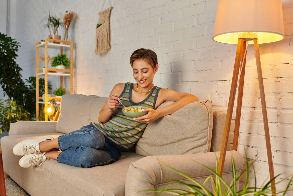 delighted veggie woman sitting on couch near lamp and eating fresh fruit salad on cozy sofa - Photo, Image