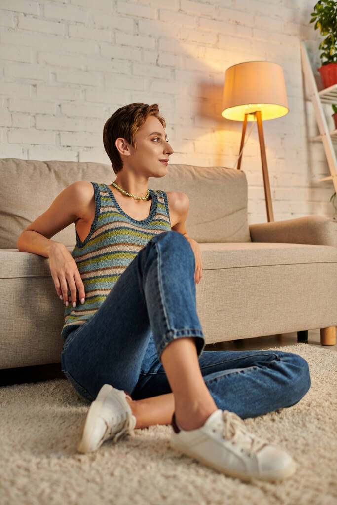 позитивна і мрійлива молода жінка, дивлячись далеко, сидячи на підлозі біля дивана у вітальні - Фото, зображення