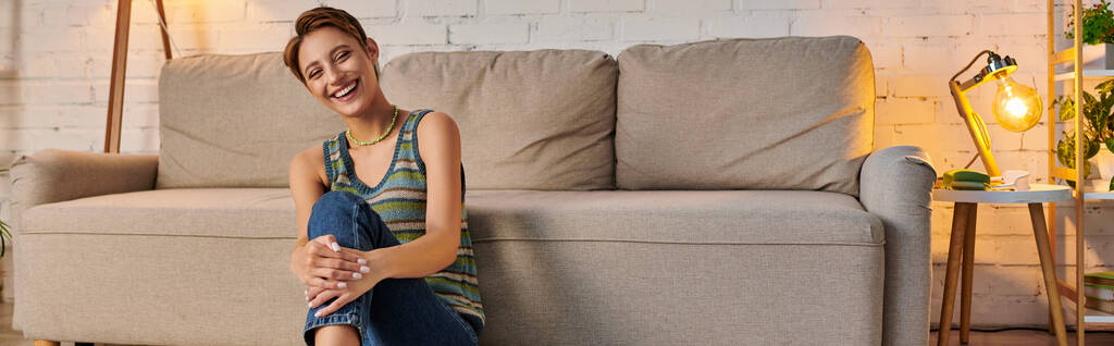 radosna młoda kobieta siedząca blisko kanapy w przytulnym salonie i patrząca w kamerę, baner poziomy - Zdjęcie, obraz