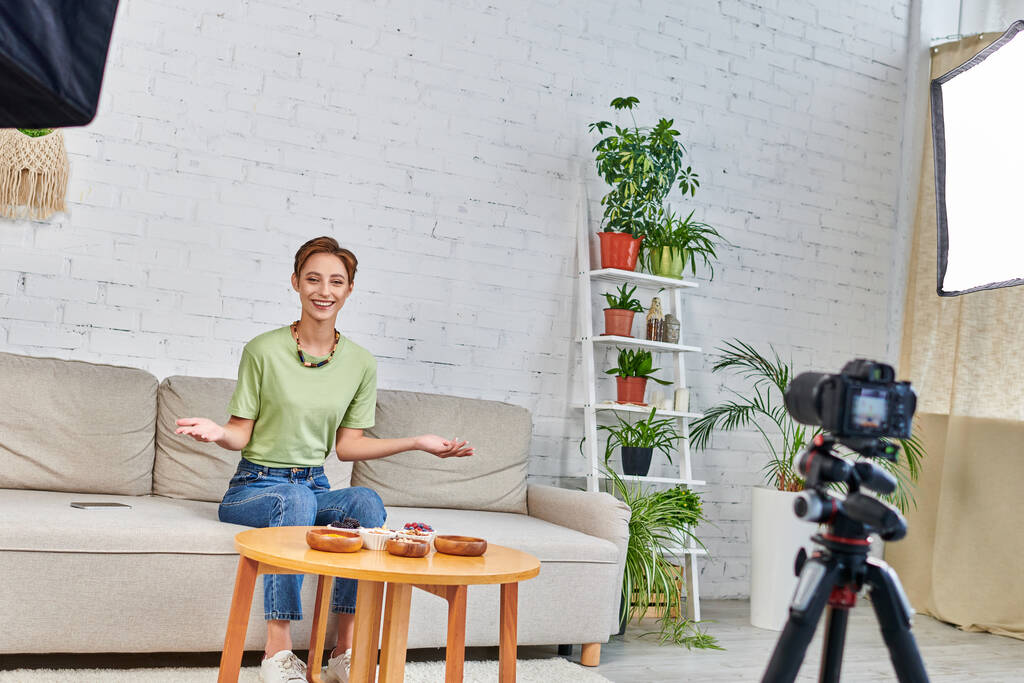 smiling woman presenting vegetarian meal during video blog on plant-based diets in green living room - Photo, Image