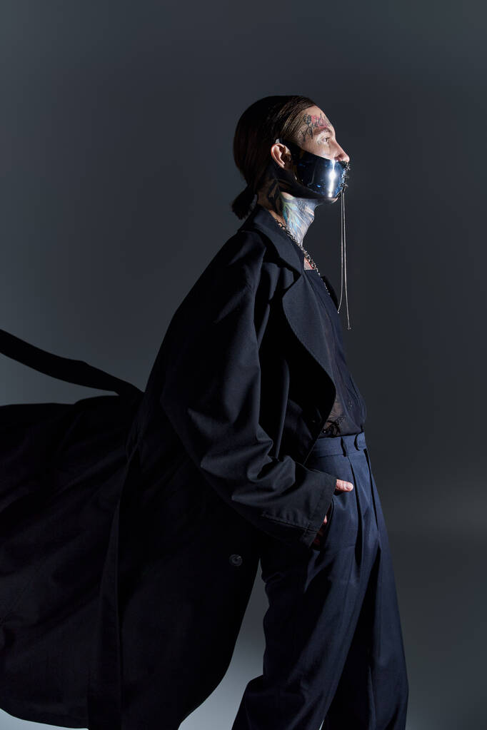 beau modèle masculin en manteau noir et masque laqué futuriste posant de profil avec les mains dans les poches - Photo, image