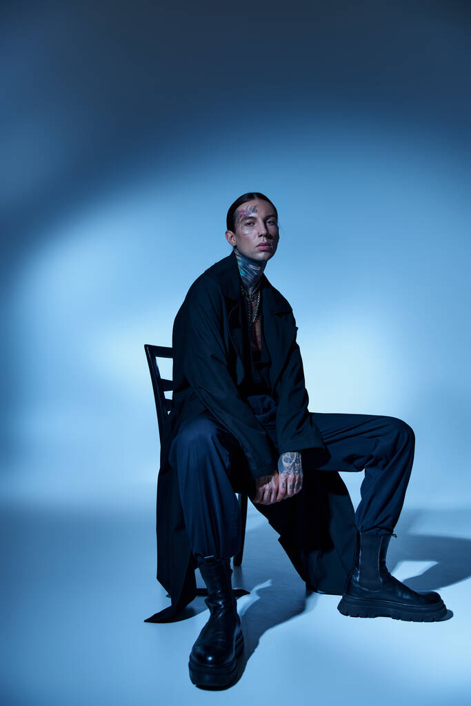 przystojny stylowy mężczyzna w czarny voguish strój siedzi na krześle i patrząc w aparat, koncepcja mody - Zdjęcie, obraz