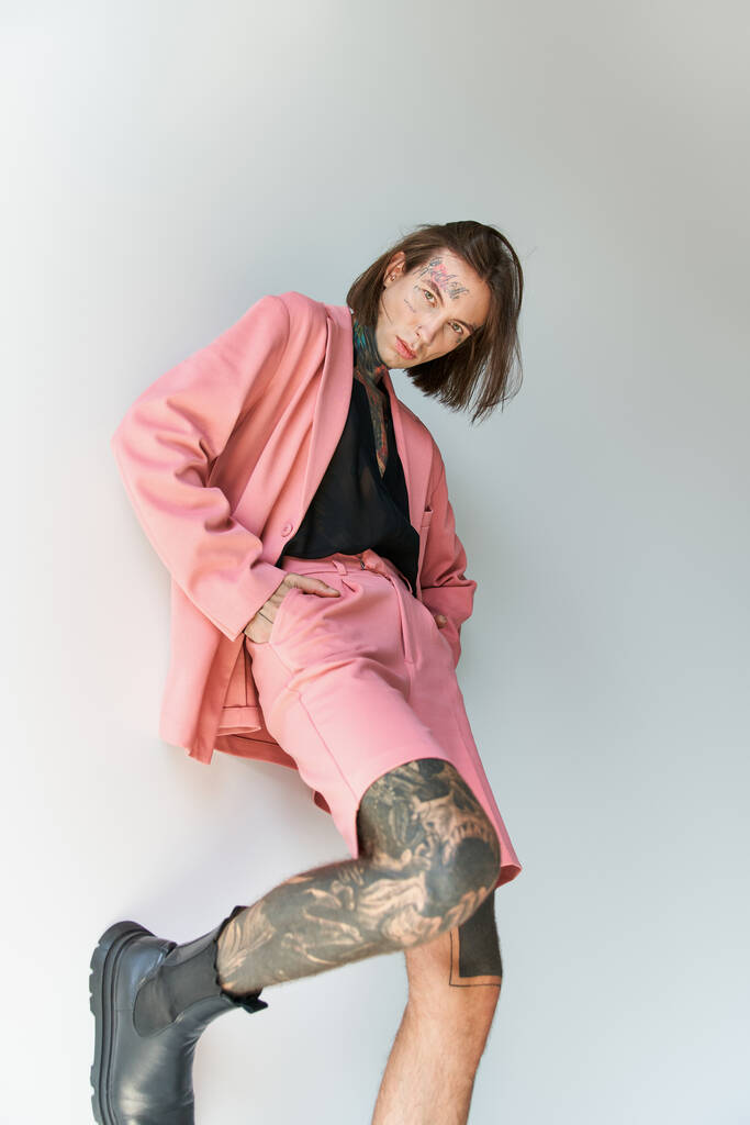 hombre atractivo de buen aspecto con tatuajes en traje elegante posando con la pierna en la pared y las manos en los bolsillos - Foto, imagen