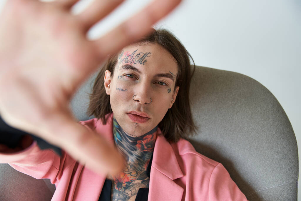 κομψό νεαρός άνδρας με τατουάζ και σκουλαρίκια σε ροζ σακάκι με το χέρι μπροστά από την κάμερα, μόδα - Φωτογραφία, εικόνα