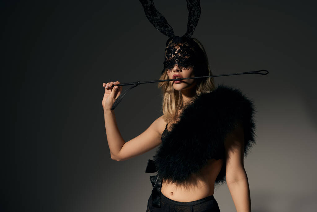 sexy joven mujer en atractivo negro traje con conejo máscara posando y sosteniendo bdsm látigo en los dientes - Foto, Imagen