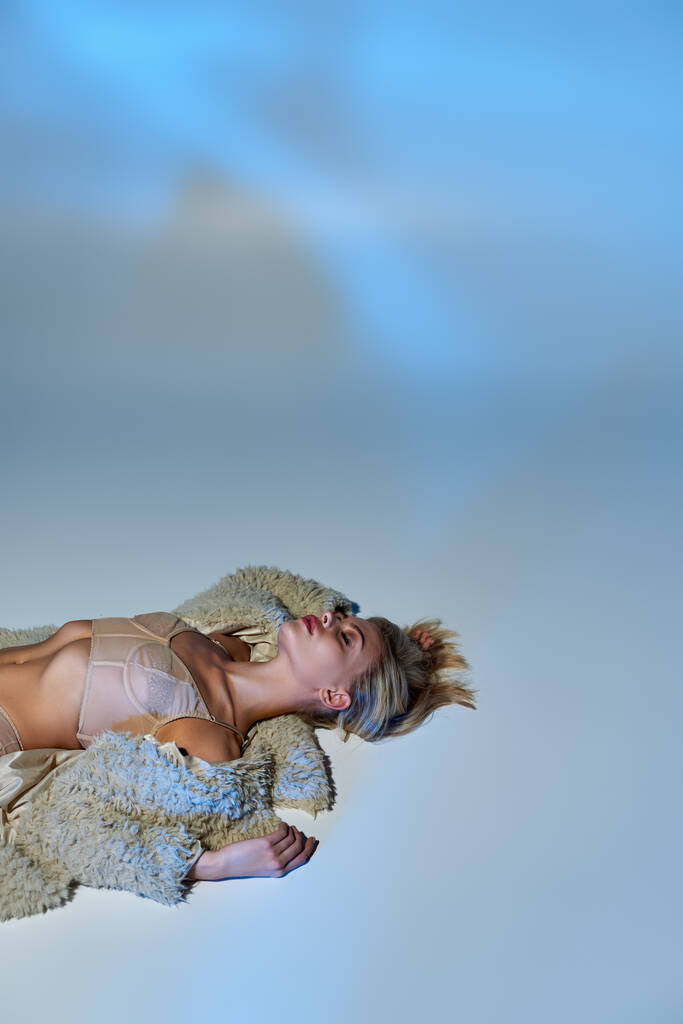 вертикальний знімок гарячої блондинки жіночої моделі в сексуальній бежевій білизні і штучному хутряному пальто лежить на підлозі - Фото, зображення