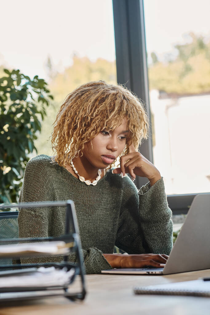 εστίασε νεαρή Αφροαμερικανή γυναίκα με σγουρά μαλλιά κοιτάζοντας το laptop της, διαδικασία εργασίας - Φωτογραφία, εικόνα