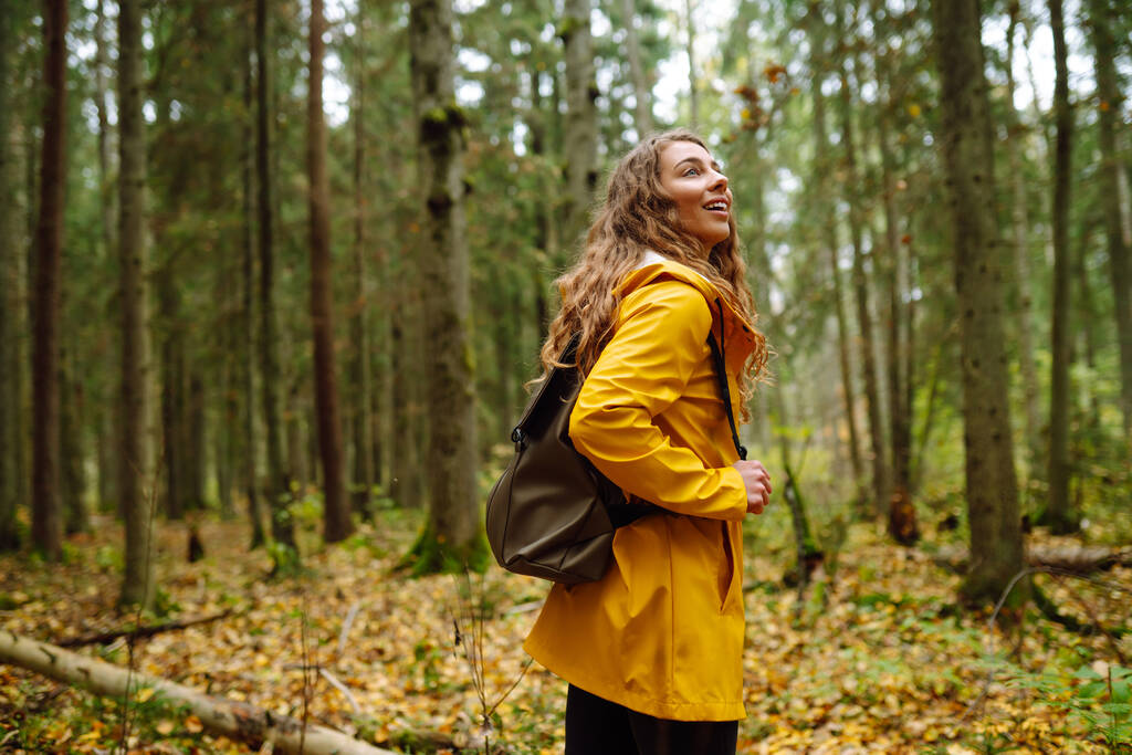 Donna felice in un cappotto giallo gode del verde bella foresta naturale intorno a lei. Un turista con uno zaino sta esplorando nuovi luoghi, passeggiando nella boscaglia della foresta. - Foto, immagini