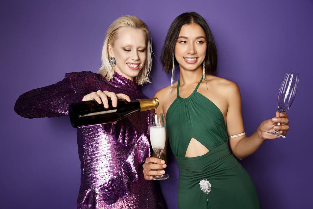 szczęśliwa kobieta wlewając szampana do szklanki azjatyckiej koleżanki na fioletowym tle, Nowy Rok 2024 - Zdjęcie, obraz