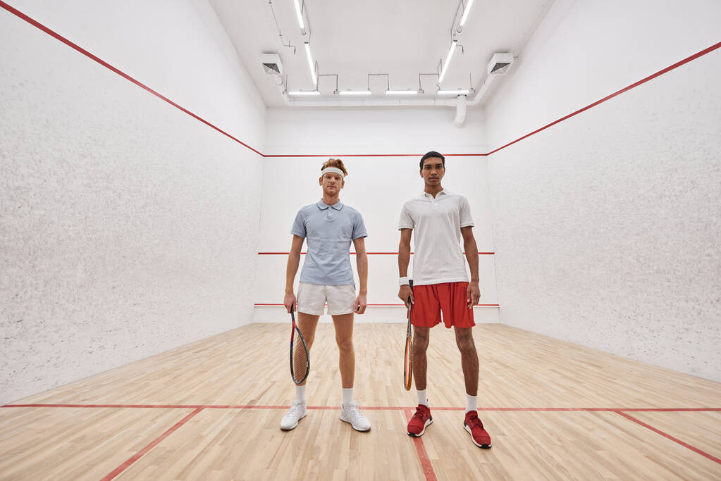 young interracial male friends in sportswear looking at camera and holding racquets for squash - Photo, Image