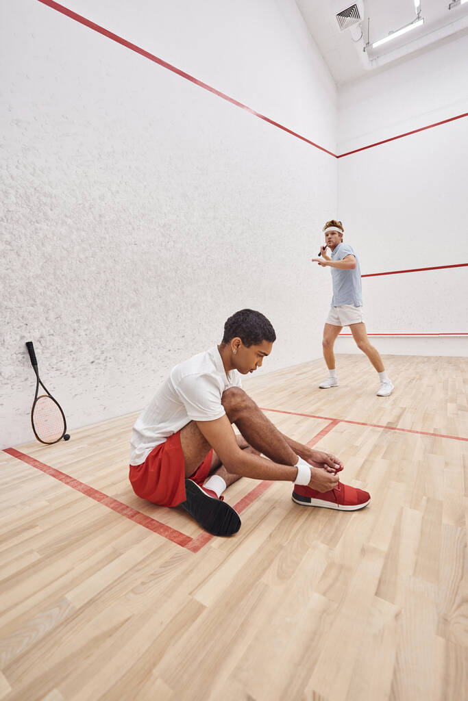 african american man tying shoelaces while sitting near redhead friend inside of squash court - Photo, Image