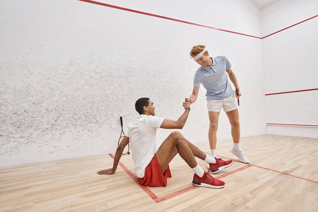 happy redhead man helping his african american friend to stand up from floor inside of squash court - Photo, Image