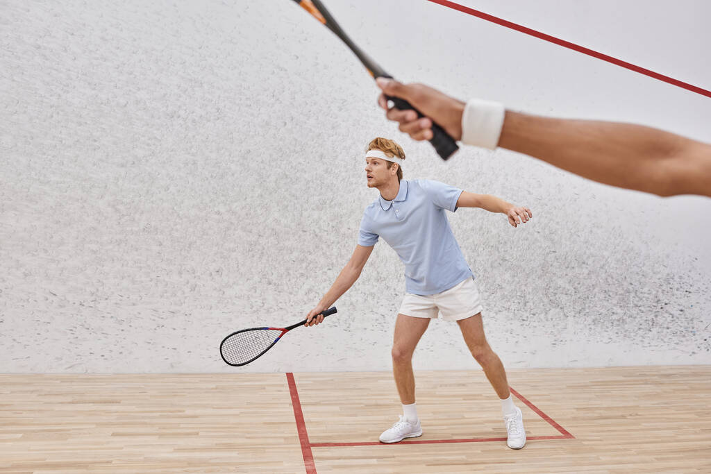 pelirrojo en ropa deportiva y diadema jugando con un amigo afroamericano dentro de la cancha de squash - Foto, Imagen